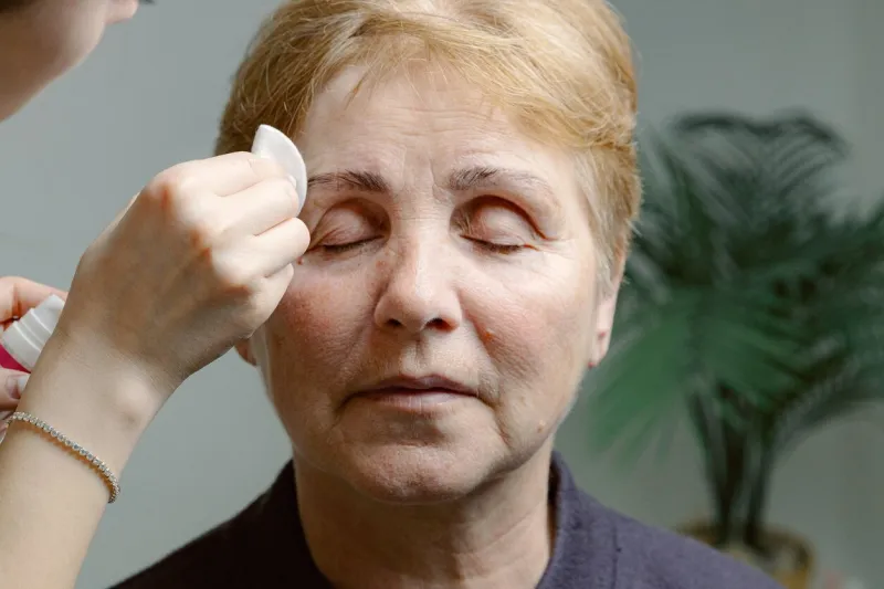 one young caucasian young unrecognizable girl cosmetologist carefully removes the remaining paint from the right eyebrow of an elderly woman with closed eyes with a cotton pad, using a cleanser, in a home beauty salon, close-up side view