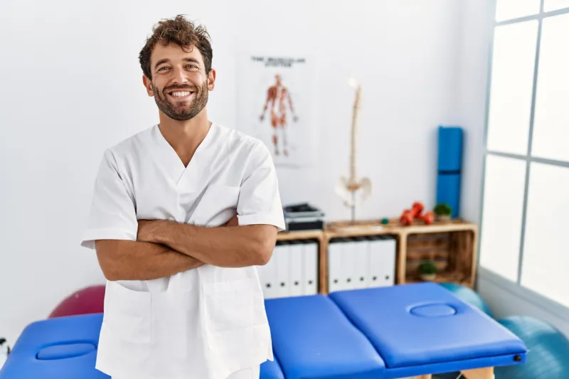 young handsome physiotherapist man working at pain recovery clinic happy face smiling with crossed arms looking at the camera positive person