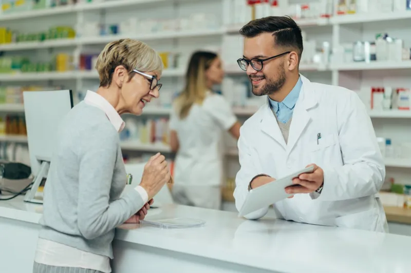 young male pharmacist giving prescription medications to senior female customer in a pharmacy with female pharmacist in the background