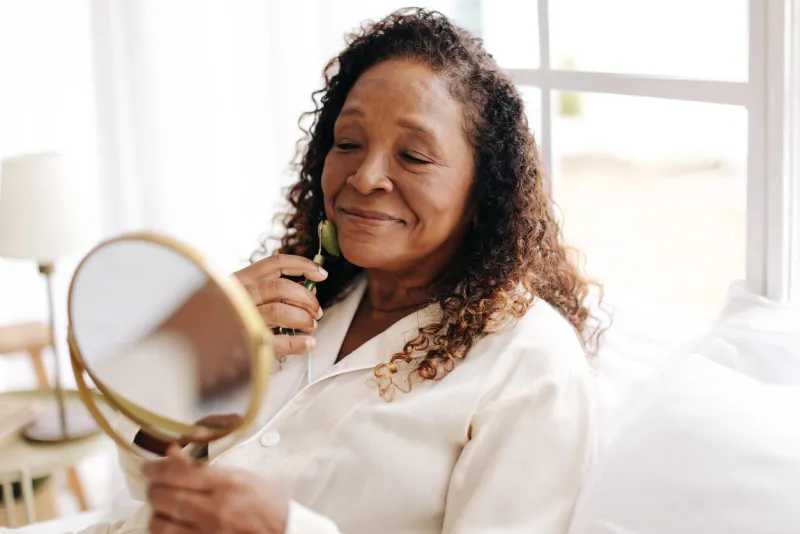 mature black woman taking care of her skin with an anti-aging jade roller, looking herself in the mirror as she rolls the cool stone over her face to promote circulation and reduce puffiness