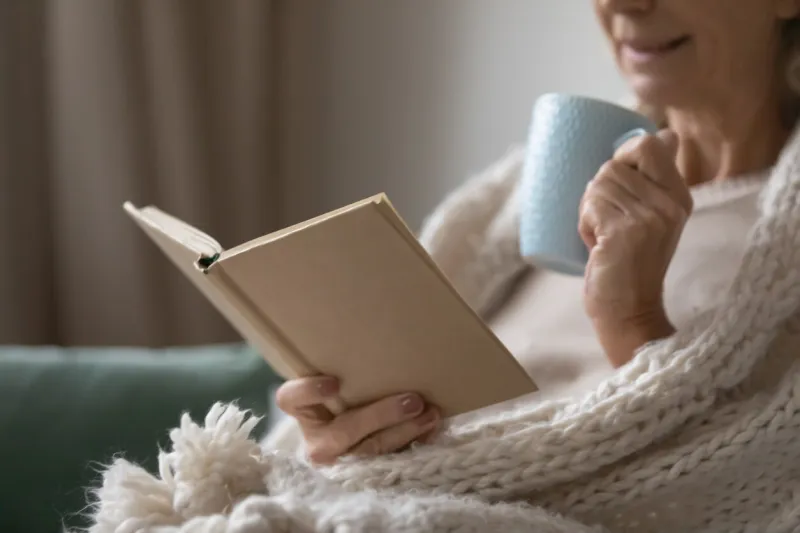 senior elderly lady wrapped in woolen comforter reading learning book, getting knowledge over cup of hot drink mature woman enjoying leisure on couch, morning coffee, studying at home close up