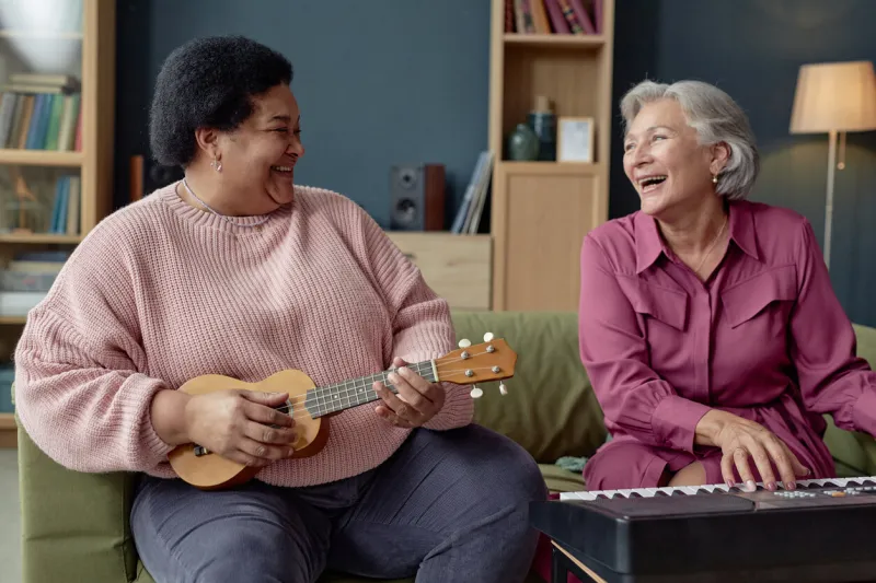 portrait of two senior women laughing and playing music together looking at each other with piano and ukulele