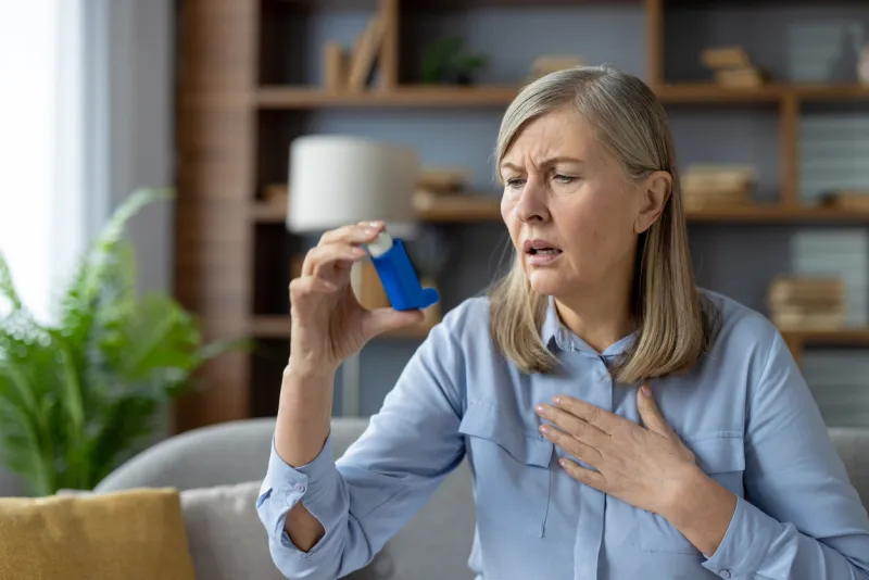 an elderly woman experiences an asthma attack and uses a blue inhaler she looks concerned while sitting in a modern living room