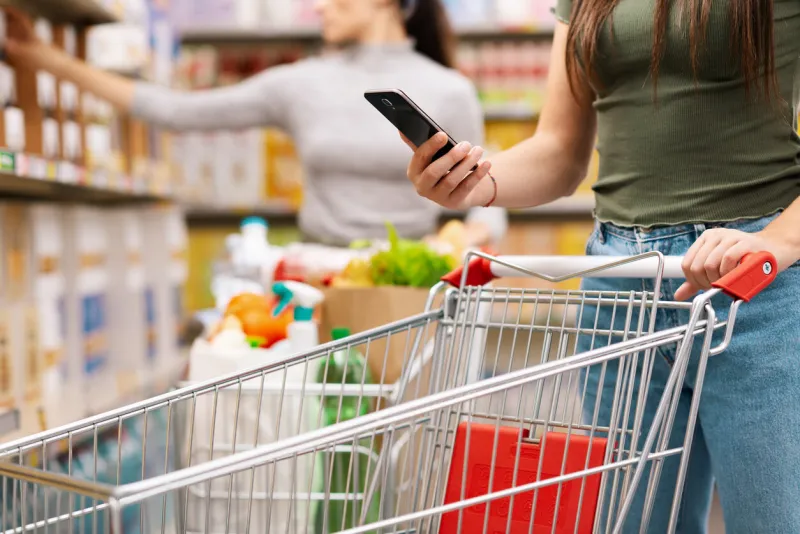 woman buying groceries at the supermarket, she is pushing a shopping cart and using her smartphone, unrecognizable people