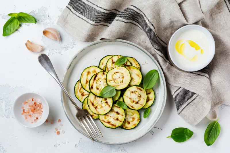 grilled zucchini salad with basil leaves, yogurt sauce and fried bread in a simple ceramic plate on a white concrete background flat lay with copy space