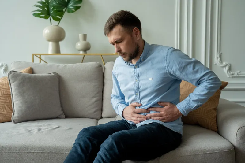 a man suffers from abdominal pain while sitting at home on the couch beautiful young man suffering from abdominal pain sitting on the couch at home