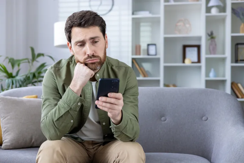 thoughtful man sitting on sofa looking at smartphone at home pensive expression, appears deep in thought or concerned casual indoor setting with focus on man's face and phone