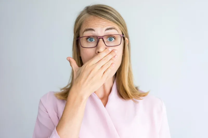 close-up of shocked young caucasian businesswoman wearing pink blouse and glasses covering mouth with hand surprise and confusion concept