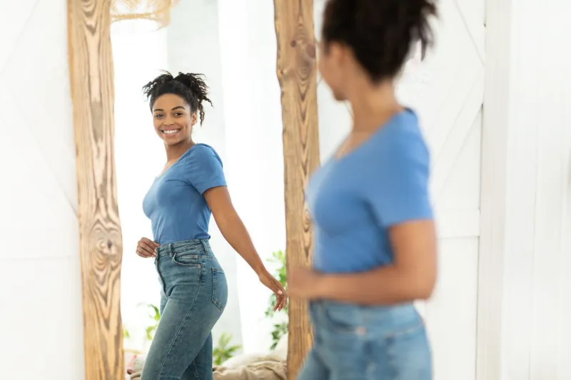 successful weight loss joyful african american girl after slimming smiling to her reflection in mirror standing at home staying fit, dieting and weight-loss concept selective focus