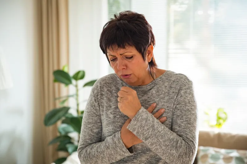 sick mature woman with sore throat, standing in living room at home catching cold, having cough