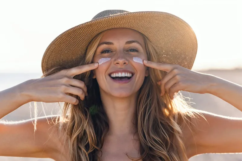 portrait of beautiful smiling woman applying sunscreen on her face while looking at camera at the beach