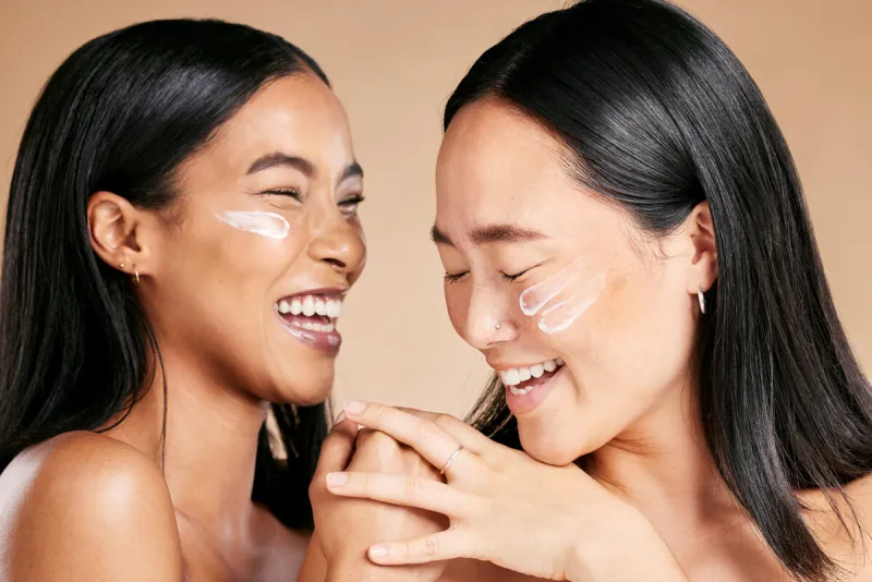 happy, beauty and skincare women with cream for face isolated on a beige studio background wellness, smile and asian model friends with sunscreen for love of cosmetics and dermatology on a backdrop