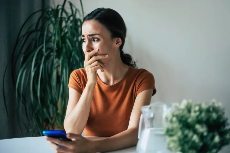sad, frustrated young brunette woman is crying with smartphone in hands while she sitting on the chair at apartment