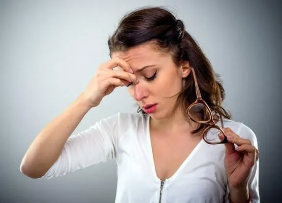 séduisante jeune femme avec un mal de tête tenant ses lunettes dans une main comme elle pince le pont de son nez avec une expression peinée, la tête et les épaules sur gris