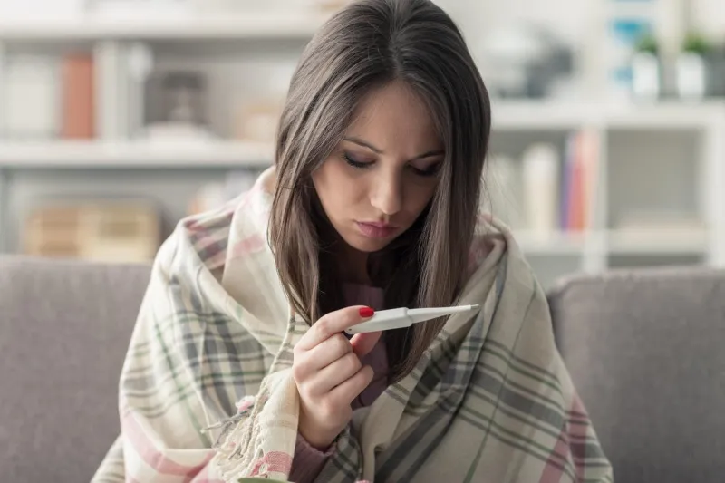 sick young woman at home on the sofa, she is covering with a blanket and taking temperature with a thermometer