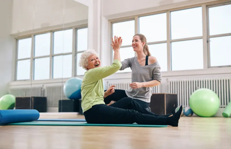 senior woman sitting on fitness mat giving high five to her personal trainer at gym excited old woman rejoicing health success with her instructor at rehab