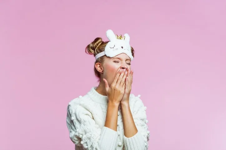 portrait of teenager wearing white sweater and cat blindfold sleep mask on her forehead, yawning with eyes closed studio shot on pink background