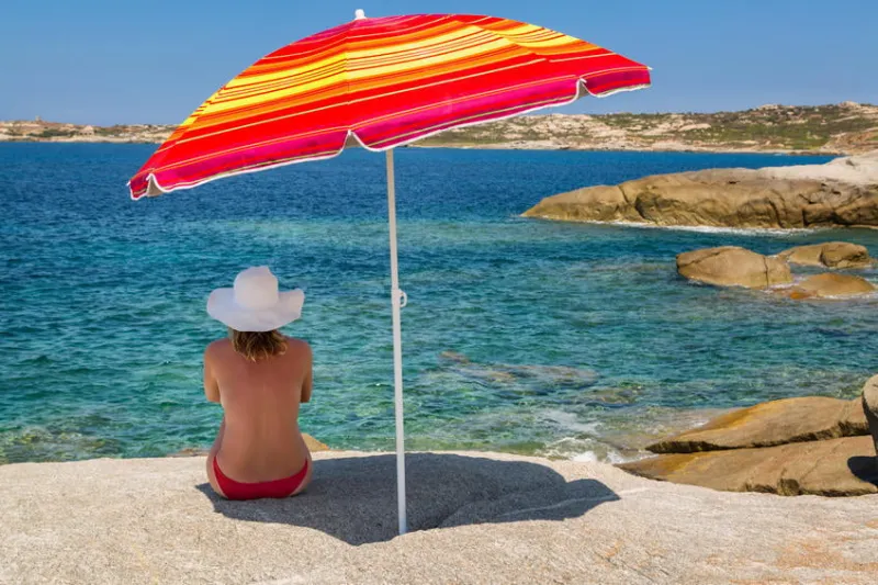 femme en bikini rouge et grand chapeau blanc assis sous un parasol rayé rouge et jaune aux couleurs vives sur un rocher et vue sur la mer méditerranée