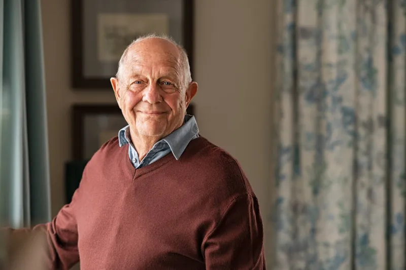 portrait of happy retired senior man standing at home near window satisfied old man in casual clothes looking at camera and smiling while standing near the window positive and confident elderly enjoy his retirement at care facility