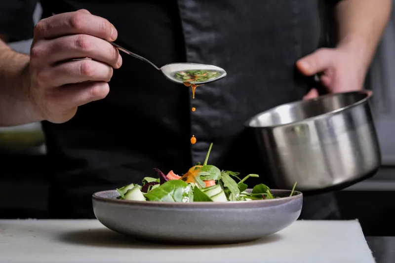 close-up of the hands of a male chef on a black background pour sauce from the spoon on the salad dish food decoration
