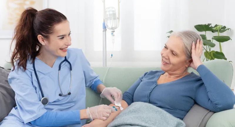 young smiling nurse or doctor help sick senior elderly woman patient in receiving iv drip infusion and vitamin injection therapy sitting on sofa in medical clinic or at home health care concept