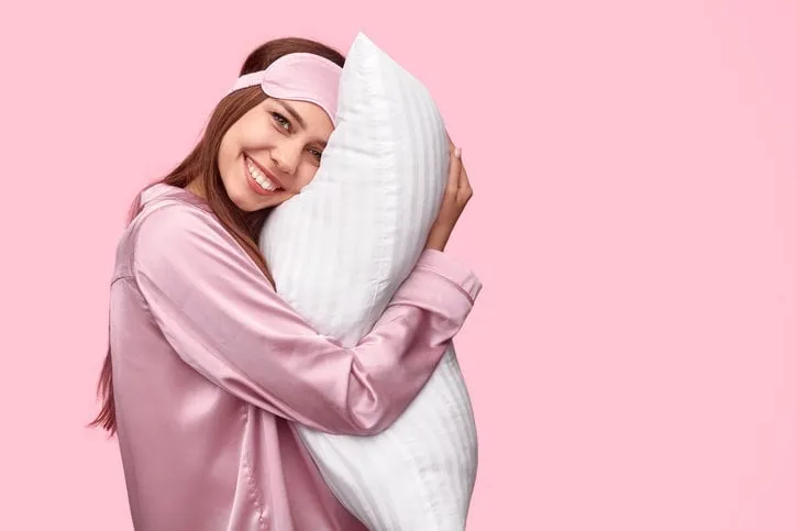 side view of delighted young woman in nightwear smiling and embracing soft pillow after good night sleep against pink background