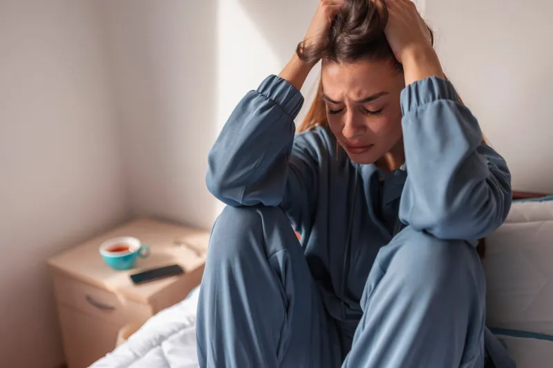 anxious young woman wearing pajamas, sitting on bed in the morning, holding head in hands, crying