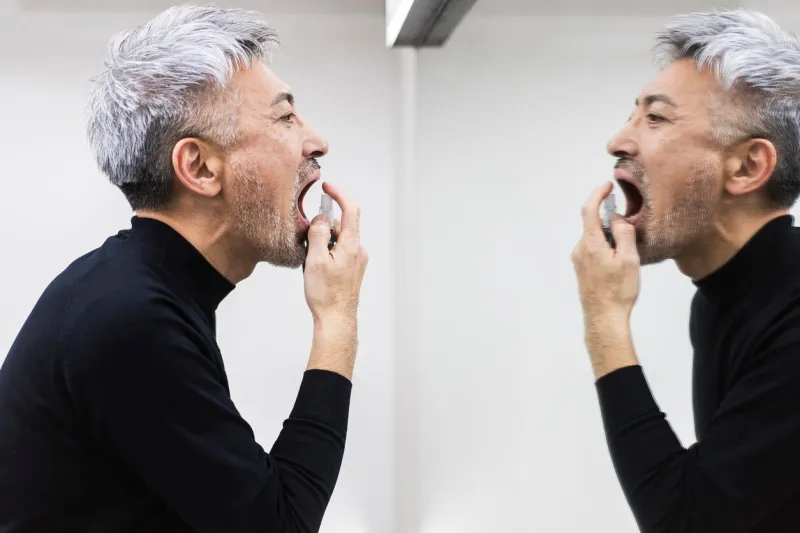 a middle-aged man uses mouth spray to treat bad breath in front of the mirror