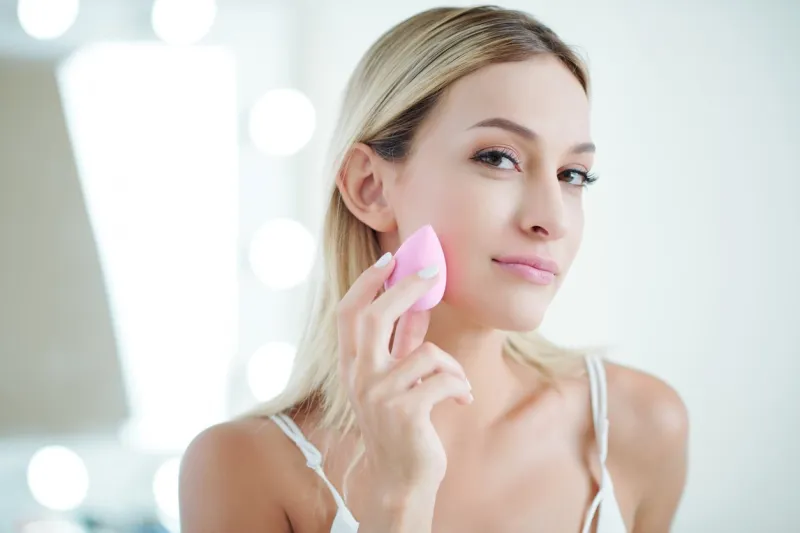 pretty young caucasian woman doing make-up and applying foundation on her skin