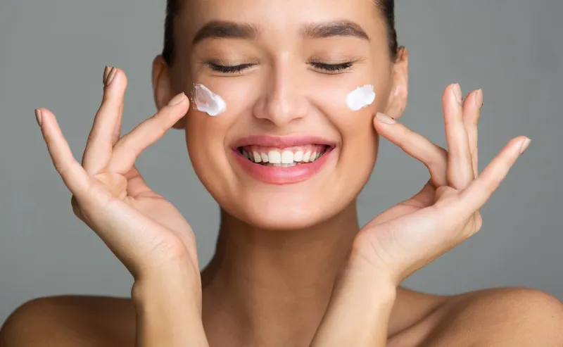 skin care happy woman applying cosmetic cream on her face on grey background