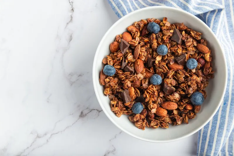 homemade chocolate granola, muesli with almonds, hazelnuts and blueberries in bowl on white marble background healthy breakfast top view flat lay minimalist design space for text