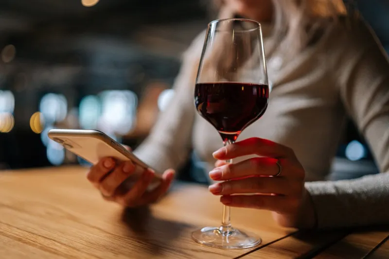 close-up cropped shot of unrecognizable young woman using smartphone, typing online message sitting at table holding in hand glass of red wine at restaurant cute lady chatting with boyfriend