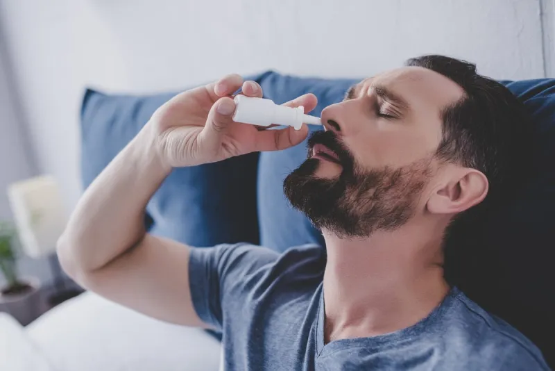 portrait of man sitting in the bed spraying drops in his nose