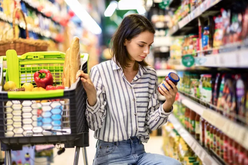 consumption and consumerism portrait of young woman with shopping cart in market buying groceries food taking products from shelves in store, holding glass jar of sauce, checking label or expiry date