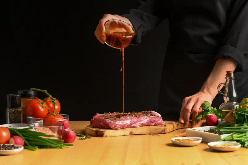 chef cooks pork steak pouring sauce, marinade on a background with vegetables recipe book, cooking, restaurant business