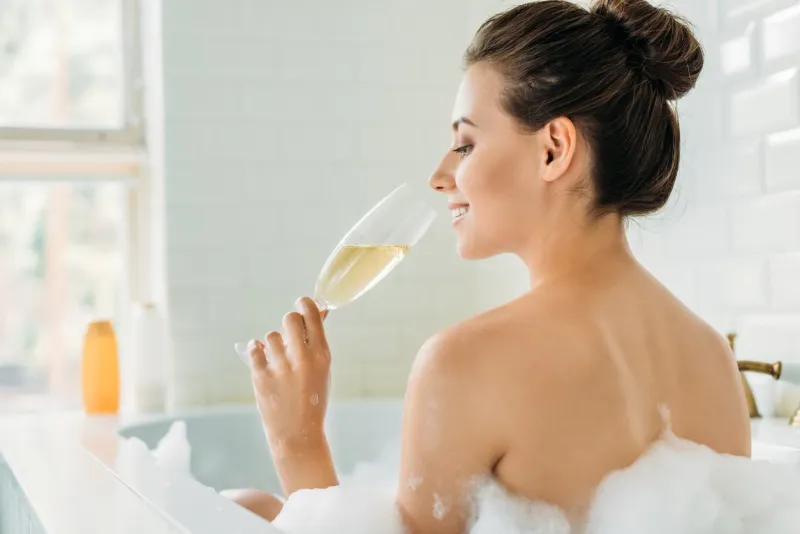rear view of beautiful smiling girl drinking wine in bathtub with foam