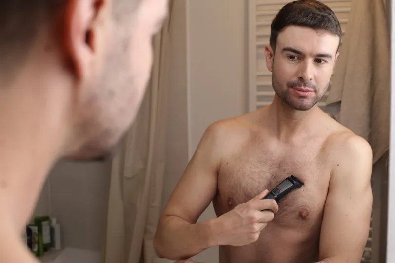 a muscular and handsome man in his forties is trimming his chest hair he looks at his mirror reflection while doing it