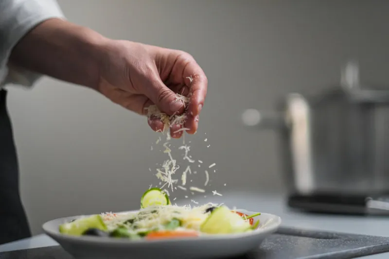 a female chef in a white uniform and a black apron in the restaurant kitchen cooking the cook sprinkles grated cheese prepared food caesar salad