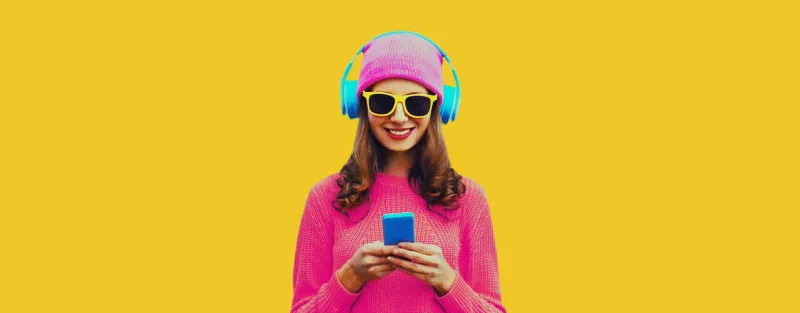 portrait of modern young woman in wireless headphones listening to music with smartphone wearing knitted sweater, pink hat on yellow background