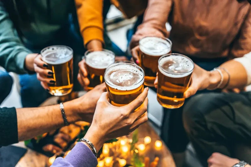 group of friends drinking and toasting glass of beer at brewery pub restaurant- happy multiracial people enjoying happy hour with pint sitting at bar table- youth food and beverage lifestyle concept