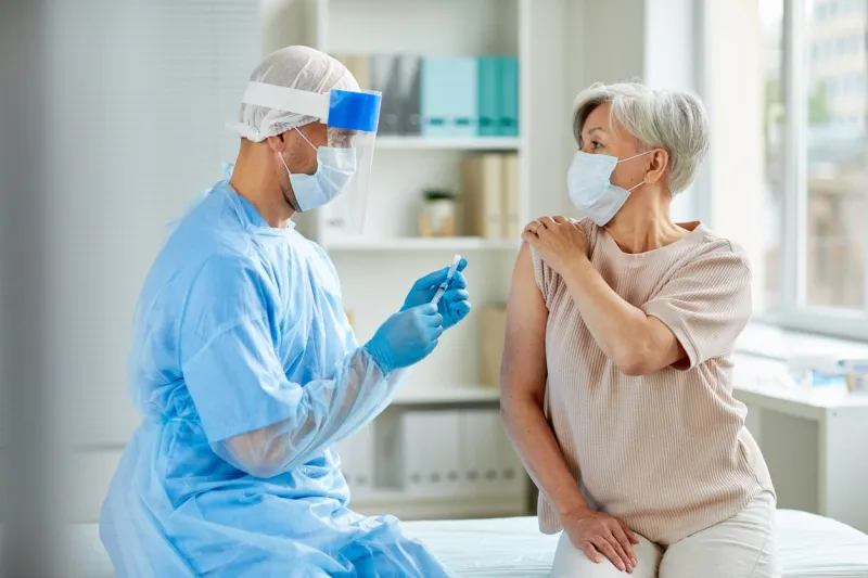 side view shot of male nurse wearing protective mask and gloves preparing medical syringe for giving injection to senior patient