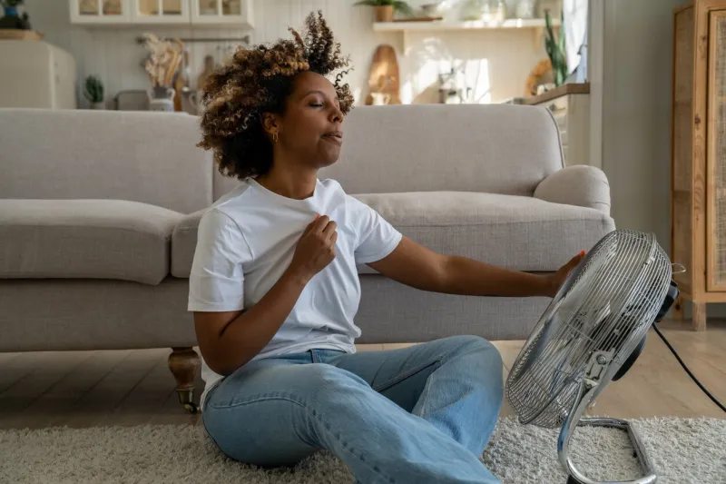 summer heat young african american woman cooling down by ventilator at home, feeling unwell with high temperature during hot weather, sitting on floor in front of electric fan during extreme heatwave