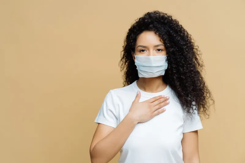 serious afro american woman wears medical face mask, has problems with breathing, presses hand to chest, got infected with coronavirus, isolated on beige ackground covid 19, health care concept