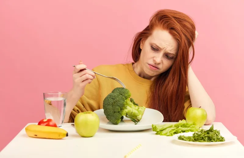 studio portrait of upset stressed dull discontent unsatisfied displeased red-haired woman holding fork in hand looking at fresh broccoli as if it is smth not tasty unappetizing