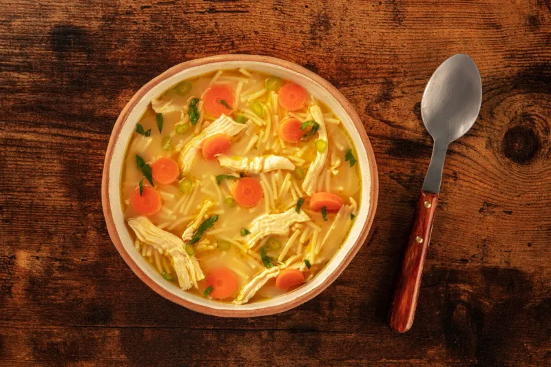 chicken noodle soup with vegetables, a bowl of healthy stock with a spoon, top shot on a rustic wooden table, winter home cooking