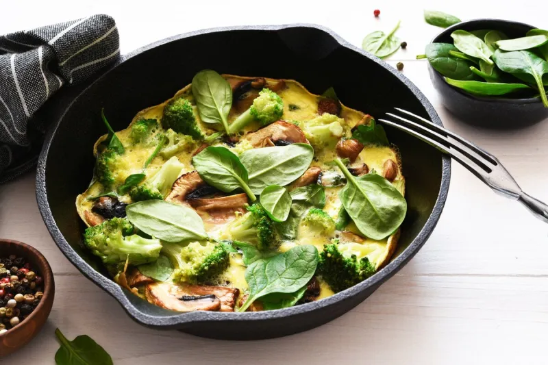 omelet with spinach and mushrooms in a cast-iron pan on a light wood background fritata with spinach