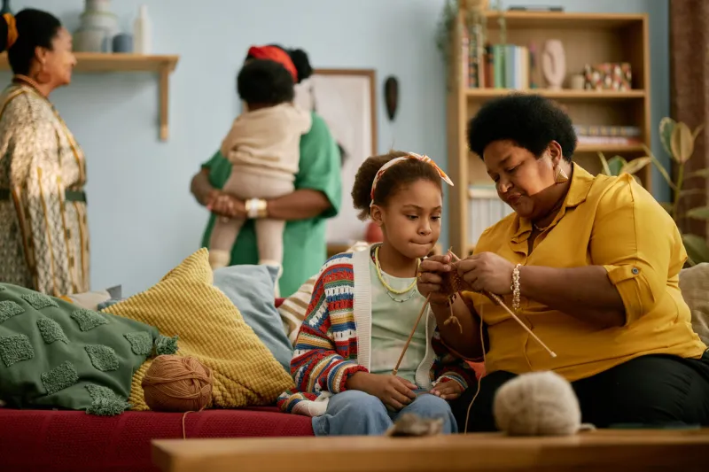 african american senior woman teaching child knitting skills while sitting on couch together during family gathering in living room