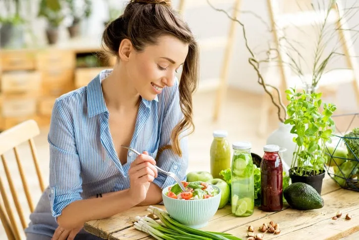 young and happy woman eating healthy salad sitting on the table with green fresh ingredients indoors
