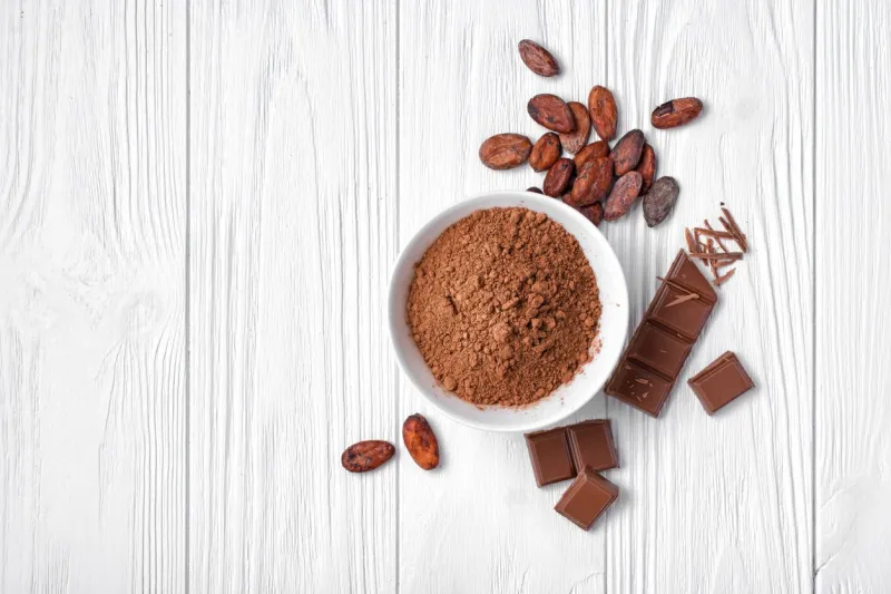 top view of cocoa powder with broken chocolate bar and cocoa beans for confectionery on white wooden background with copy space