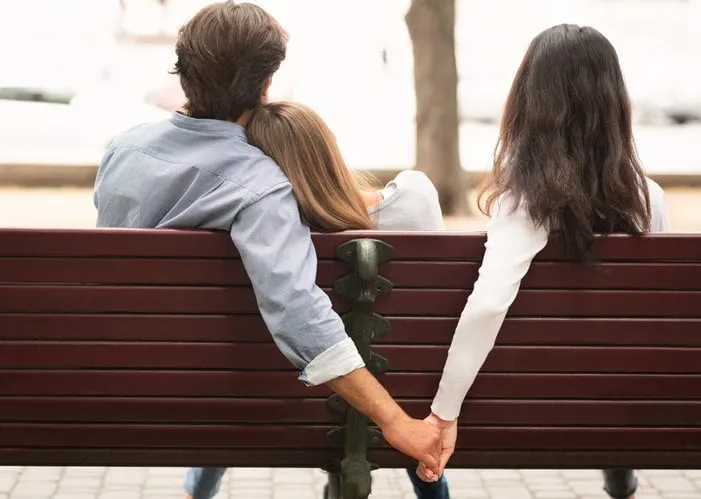 love triangle cheating boyfriend hugging girlfriend holding hands with her girl friend sitting on bench together in park outdoor back-view